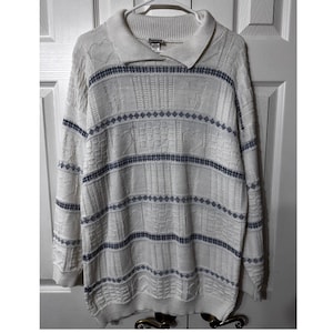 May include: A cream-coloured sweater with a collared neckline and long sleeves. The sweater features textured patterns and horizontal stripes in navy blue. The garment is hanging on a white hanger against a white door.