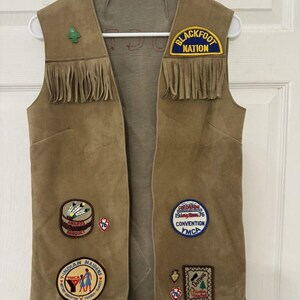 May include: Tan suede vest with fringe and patches. Patches include "Blackfoot Nation", "Awards Banquet", "Indian Maidens", and "1976 Convention YMCA". The vest has a light brown interior and is hanging on a white hanger.