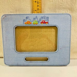 Può includere: Una cornice rettangolare azzurra con un bordo cucito. La parte superiore della cornice presenta un paesaggio urbano ricamato con tre auto colorate. La cornice ha un'apertura rettangolare per una foto e una piccola fessura nella parte inferiore.