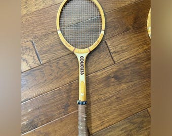 May include: A vintage wooden tennis racket with a light brown frame and a tightly strung net. The handle is wrapped in a worn, light brown grip. The racket features the brand name "YONEX" and the word "COUGUSS" printed on the handle.