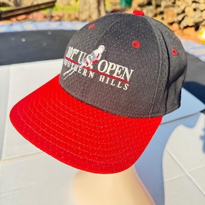 Op de afbeelding: Een zwart-rode baseballcap met de tekst "101st U.S. OPEN SOUTHERN HILLS" in het wit. De cap heeft een rode klep en rode accenten aan de bovenkant. De cap is gemaakt van mesh-materiaal.
