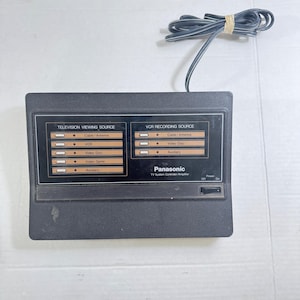 Könnte beinhalten: Ein schwarzer Panasonic TV System Controller/Amplifier mit einem Netzschalter und einem Kabel. Der Controller hat Abschnitte für Fernseh- und VCR-Aufnahmequellen, einschließlich Kabel/Antenne, VCR, Video Disc, Videospiel und Auxiliary.