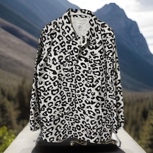 May include: A white and black leopard print jacket with a collared neckline, button-down front, and two chest pockets. The jacket is hanging on a white hanger. The background features a blurred mountain and forest scene.
