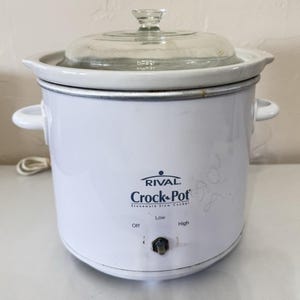 May include: White Rival Crock-Pot stoneware slow cooker with a glass lid. The slow cooker has a black control knob with settings for Off, Low, and High. The Crock-Pot has two white handles and a power cord.