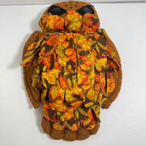 Op de afbeelding: Een handgemaakte uilversiering met een bruin lichaam en vleugels. De borst en kop van de uil zijn versierd met stof met een kleurrijk herfstbladpatroon in tinten oranje, geel en bruin. Zwarte stof wordt gebruikt voor de ogen.
