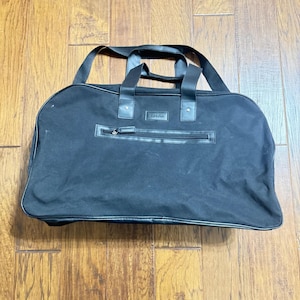 Pode incluir: Uma mala de viagem preta com formato retangular, com um bolso frontal com zíper e duas alças superiores. A mala tem uma alça de ombro e detalhes em couro. A mala está sobre uma superfície de madeira.