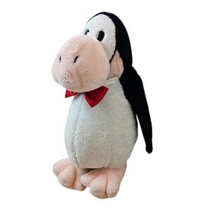 Pode incluir: Um brinquedo de pelúcia de pinguim com corpo branco, asas e cabeça pretas e um grande bico rosa. Possui uma gravata borboleta vermelha e pés rosa. O pinguim está em pé e parece macio e fofinho.
