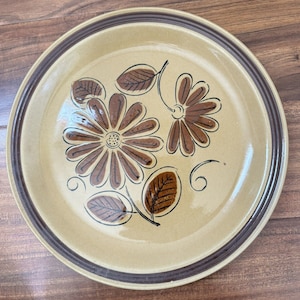 以下が含まれることがあります： 茶色の花の模様が入った、ベージュ色のセラミックプレート。 プレートには、黒いアウトラインの2つの茶色の花と葉が描かれています。 茶色の二重のボーダーがプレートの端を縁取っています。 プレートは木製の表面にあります。