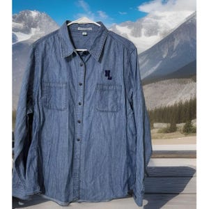 May include: A long-sleeved denim shirt in a medium blue wash, featuring a button-down front, two chest pockets, and a classic collar. The shirt has the embroidered initials "NL" in navy blue on the left chest. The shirt is hanging on a white hanger.