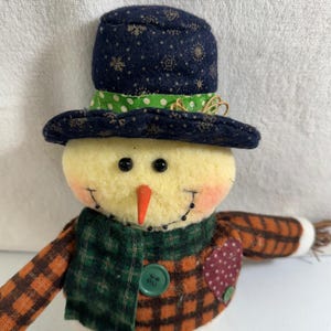 Può includere: Un pupazzo di neve fatto a mano con un cappello a cilindro blu navy adornato con fiocchi di neve dorati e una fascia verde a pois. Il pupazzo di neve ha un naso a carota, occhi a bottone neri e una sciarpa e una giacca a quadri. Una decorazione invernale festosa.