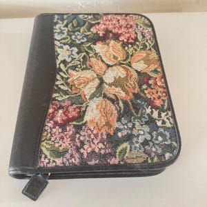 May include: A dark brown zippered case with a floral tapestry design. The tapestry features a variety of flowers in shades of pink, orange, and blue. The case has a leather-like appearance.