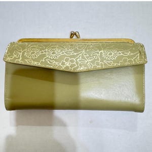 May include: A sage green wallet with a gold-colored clasp and floral design. The top flap features a gold floral pattern, while the body of the wallet is a solid, muted green. The clasp is a classic kiss-lock style.