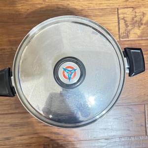 May include: Stainless steel pot with a lid. The lid features a circular emblem with the text "UNITED STATES" and a stylized "S" within a blue and red design. The pot has black handles.