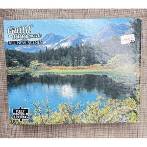 May include: A Guild 1000-piece jigsaw puzzle depicting a landscape with a lake, mountains, and trees. The box reads "ALL NEW SCENES" and "FULLY INTERLOCKING". The image shows a tranquil outdoor scene with a blue lake and mountain range.