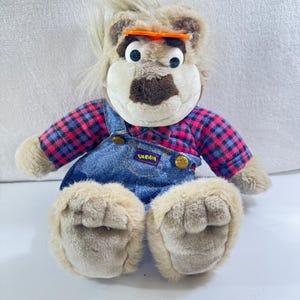 Puede incluir: Un oso de peluche con cara, orejas y patas de color marrón claro. Viste una camisa a cuadros en rosa, morado y negro, y un peto vaquero azul. El peto tiene el nombre "Qubba".