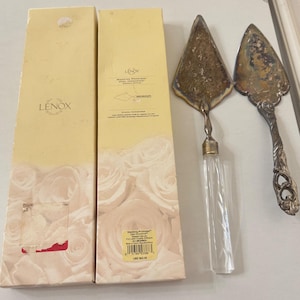 May include: Two cake servers with ornate handles and a clear glass handle. The servers are next to two boxes with the Lenox logo and the words "Wedding Promises" on them. The boxes have a floral design.