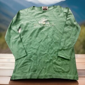 May include: A long-sleeved, green shirt with two front pockets. The shirt features an embroidered design above the text "COLORADO". The shirt is made of a soft, textured fabric and has a crew neck.