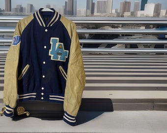 May include: A navy blue and tan varsity jacket with the letters "LA" on the front. The jacket has a white collar and cuffs with blue stripes. A patch is visible on the left sleeve. The jacket is hanging on a white hanger.