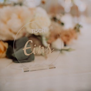 Könnte beinhalten: Eine durchsichtige Acryl-Tischnummer mit dem Text "Tisch Eins" in goldener Schrift.