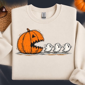Puede incluir: Una sudadera de cuello redondo color crema con un estampado gráfico de una gran calabaza naranja con la boca dentada persiguiendo a tres pequeños fantasmas blancos. El diseño está realizado en estilo boceto con contornos negros.