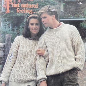 May include: Two people wearing cream-coloured knitted sweaters with cable knit patterns. The woman is wearing a brown headband and the man has short brown hair. The text "Patons" and "That natural feeling..." is visible at the top of the image. The text "Une allure naturelle" is visible at the bottom of the image.