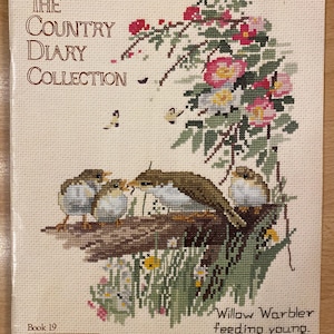 May include: A cross-stitch pattern book titled "The Country Diary Collection." The cover features a detailed cross-stitch design of a bird feeding its young, with flowers and greenery. The book is labeled "Book 19" and includes the text "Willow Warbler feeding young."