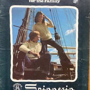 以下が含まれることがあります： 「Fisherman Fashions for the Family」と題されたヴィンテージ雑誌の表紙。クリーム色のケーブルニットセーターと濃い青色のジーンズを着た2人がヨットに乗っている様子が写っています。雑誌はSpinnerinからです。