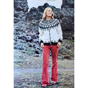 Könnte beinhalten: Eine Frau trägt einen weißen und schwarzen gemusterten Pullover mit schwarzem Knopfverschluss, eine rote Cordhose und weiße Schuhe. Sie steht vor einer felsigen Landschaft.