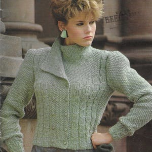 May include: A sage green knit sweater featuring a high collar, long sleeves, and a button-down front. The garment displays a textured stitch pattern with vertical cable-like details on the front and sleeves. The text 1055-514 is visible.