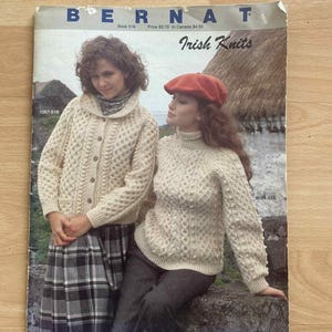 May include: Vintage "Bernat Irish Knits" book cover featuring two women in cream-colored cable-knit sweaters. One wears a beret. The book cover has the title "Irish Knits" and the Bernat logo.