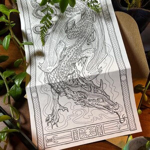 May include: Black and white dragon illustration on a folded paper. The dragon is detailed with scales, wings, and a long tongue. The paper has decorative borders and a section with symbols. The image is surrounded by greenery.