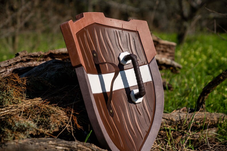May include: A brown and white wooden shield with a leather strap handle. The shield has a wood grain texture and a white stripe across the centre.