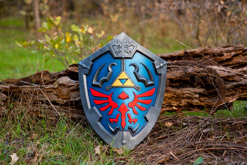 May include: A blue and silver shield with a red and yellow Triforce symbol. The shield is made of plastic and has a realistic look.