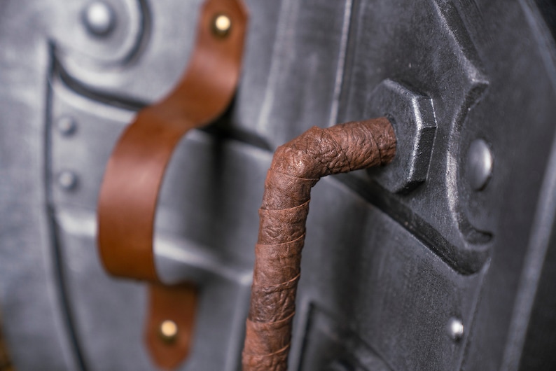 May include: Close-up of a shield with a brown leather strap and handle. The shield is made of metal and has a textured surface.