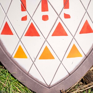 May include: A white shield with a brown border and a pattern of orange and red triangles. The shield is lying on a bed of green grass.