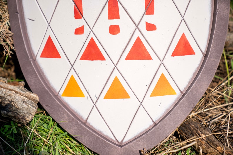 May include: A white shield with a brown border and a pattern of orange and red triangles. The shield is lying on a bed of green grass.