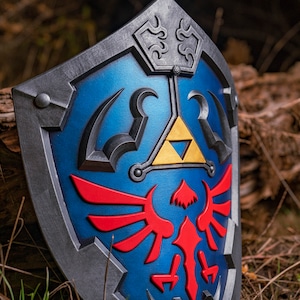 May include: A blue and silver shield with a red and gold emblem. The emblem features a stylised bird with wings spread and a triangular symbol with a gold interior.