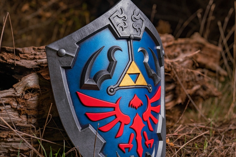 May include: A blue and silver shield with a red and gold emblem. The emblem features a stylised bird with wings spread and a triangular symbol with a gold interior.