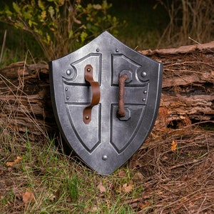 May include: A silver-coloured shield with a leather handle and metal accents. The shield is lying on a bed of brown pine needles and green grass.