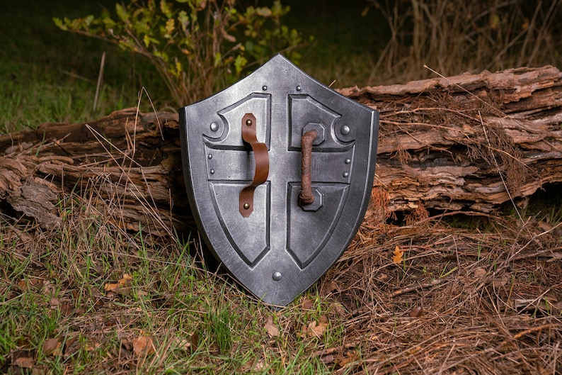 May include: A silver-coloured shield with a leather handle and metal accents. The shield is lying on a bed of brown pine needles and green grass.