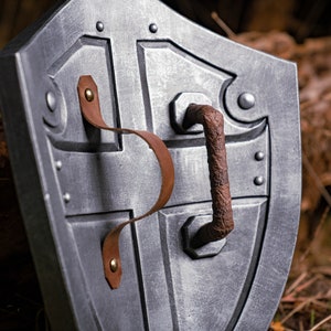 May include: A silver metal shield with a brown leather strap and a metal handle. The shield has a cross-shaped design and is decorated with rivets.