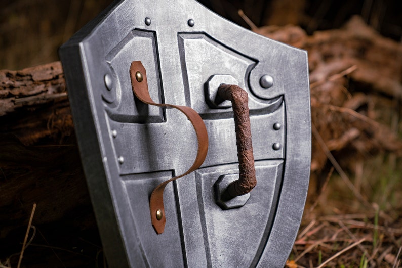May include: A silver metal shield with a brown leather strap and a metal handle. The shield has a cross-shaped design and is decorated with rivets.