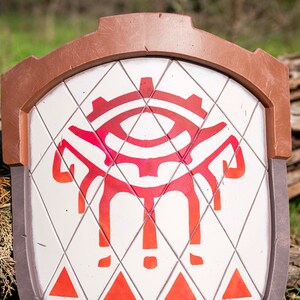 May include: A white shield with a red and orange eye design. The shield has a brown leather trim and a diamond pattern.