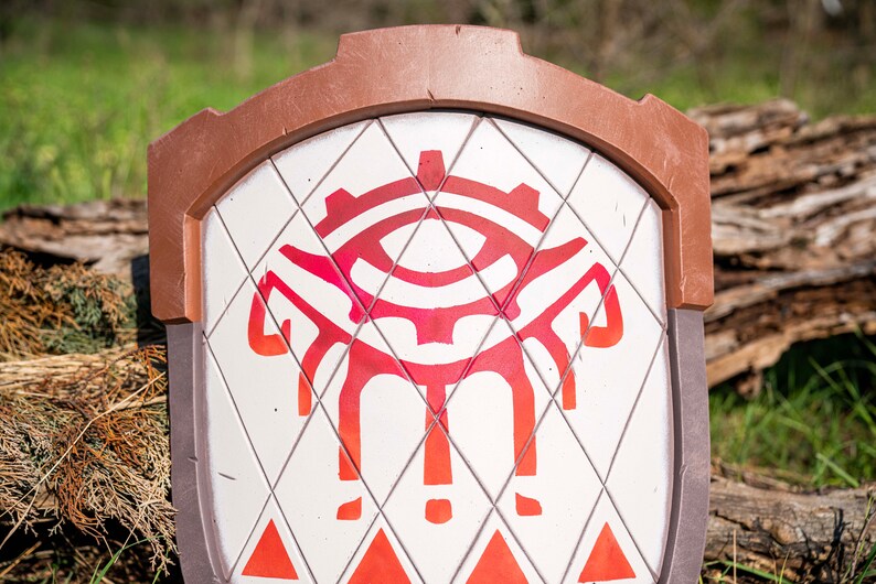 May include: A white shield with a red and orange eye design. The shield has a brown leather trim and a diamond pattern.