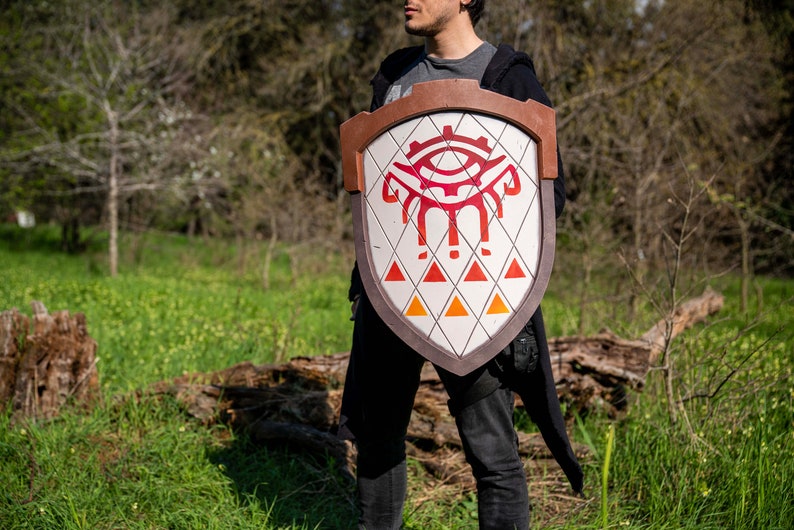 May include: A person holding a large, wooden shield with a red and white design. The shield features a stylised eye with a gear-like pattern and three triangles at the bottom.