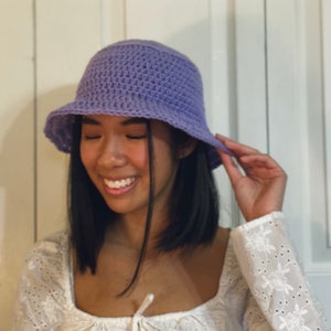 May include: A woman wearing a purple crocheted bucket hat. She is smiling and looking down. She is wearing a white, lacy top.
