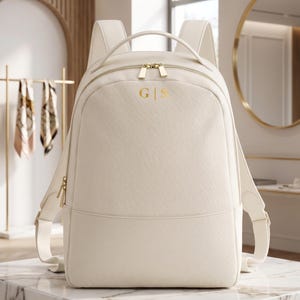 Peut inclure: Sac à dos crème avec des fermetures éclair et des accessoires dorés. Le sac à dos porte les initiales dorées "G|S" sur le devant. Le sac est fait d'un matériau texturé et possède des bretelles réglables. Le sac à dos est présenté sur une surface en marbre.