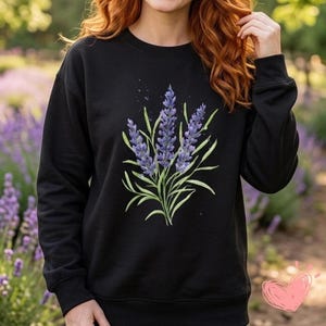 May include: A black long-sleeve sweatshirt featuring a central graphic illustration of three sprigs of purple lavender with green stems and leaves. The design includes subtle paint splatter details around the lavender flowers on the front.