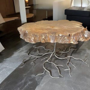 May include: A wooden coffee table with a natural, rustic finish. The table top is a thick slice of wood with a live edge. The base is made of metal and resembles tree roots.