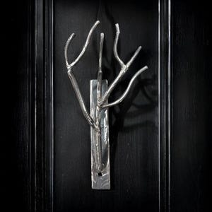 Puede incluir: Una escultura de pared de metal plateado que representa un árbol estilizado. La escultura está montada sobre una base rectangular de metal y cuelga sobre un fondo negro. Las ramas del árbol se extienden hacia afuera, creando un elemento decorativo único.